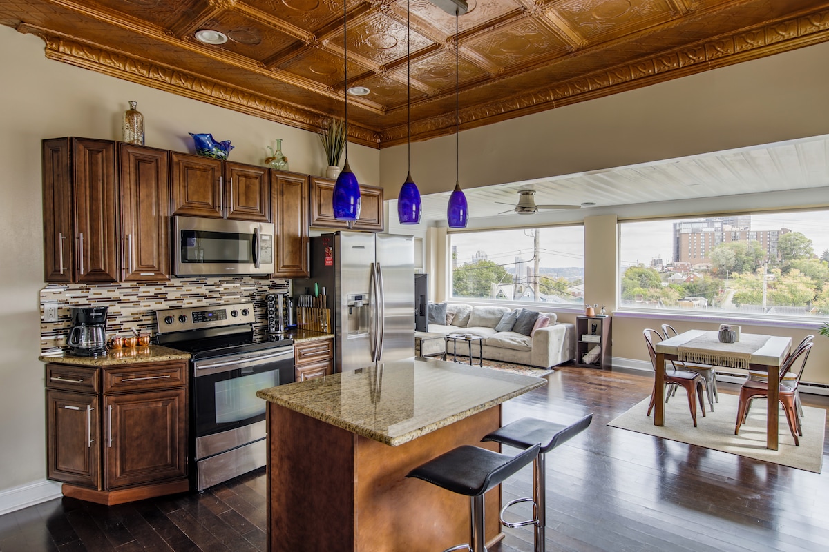 The kitchen features rich wooden cabinetry, a granite island with seating, and modern appliances. A mosaic backsplash complements the space. An open layout reveals a dining area with a large table, and the living area is visible through expansive windows, offering views of the surrounding neighborhood.