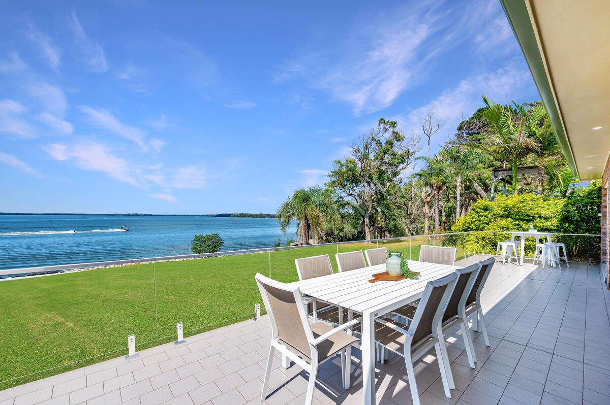 An outdoor dining area features a spacious table with light-colored chairs, set against a backdrop of lush greenery and the tranquil lake. Clear blue skies and gentle water ripples enhance the serene lakeside setting, inviting relaxation and enjoyment.