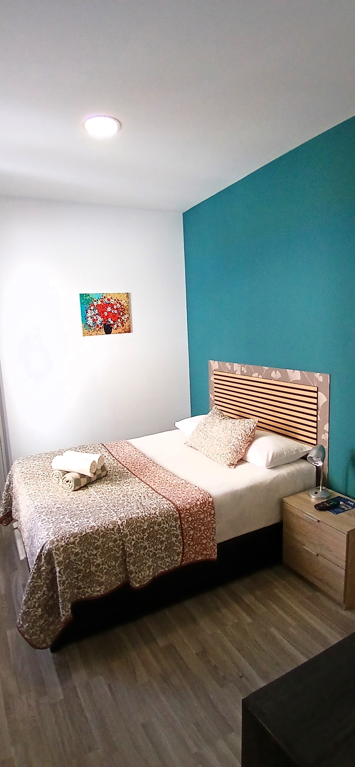 A comfortable sleeping area is highlighted by a cozy bed with a patterned blanket. A decorative wall art piece adds interest to the turquoise wall. A bedside table features a lamp and a small tray, while ample natural light fills the room.