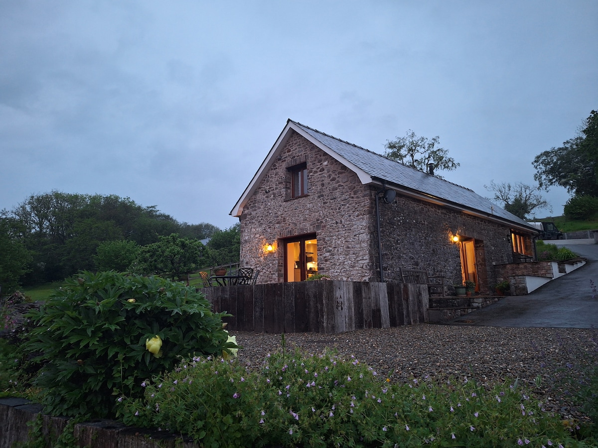 A stone barn with a sloped roof is illuminated by warm light from its windows. Surrounded by lush greenery and gravel pathways, the structure blends with the tranquil rural landscape, reflecting its serene setting in the Brecon Beacons National Park.