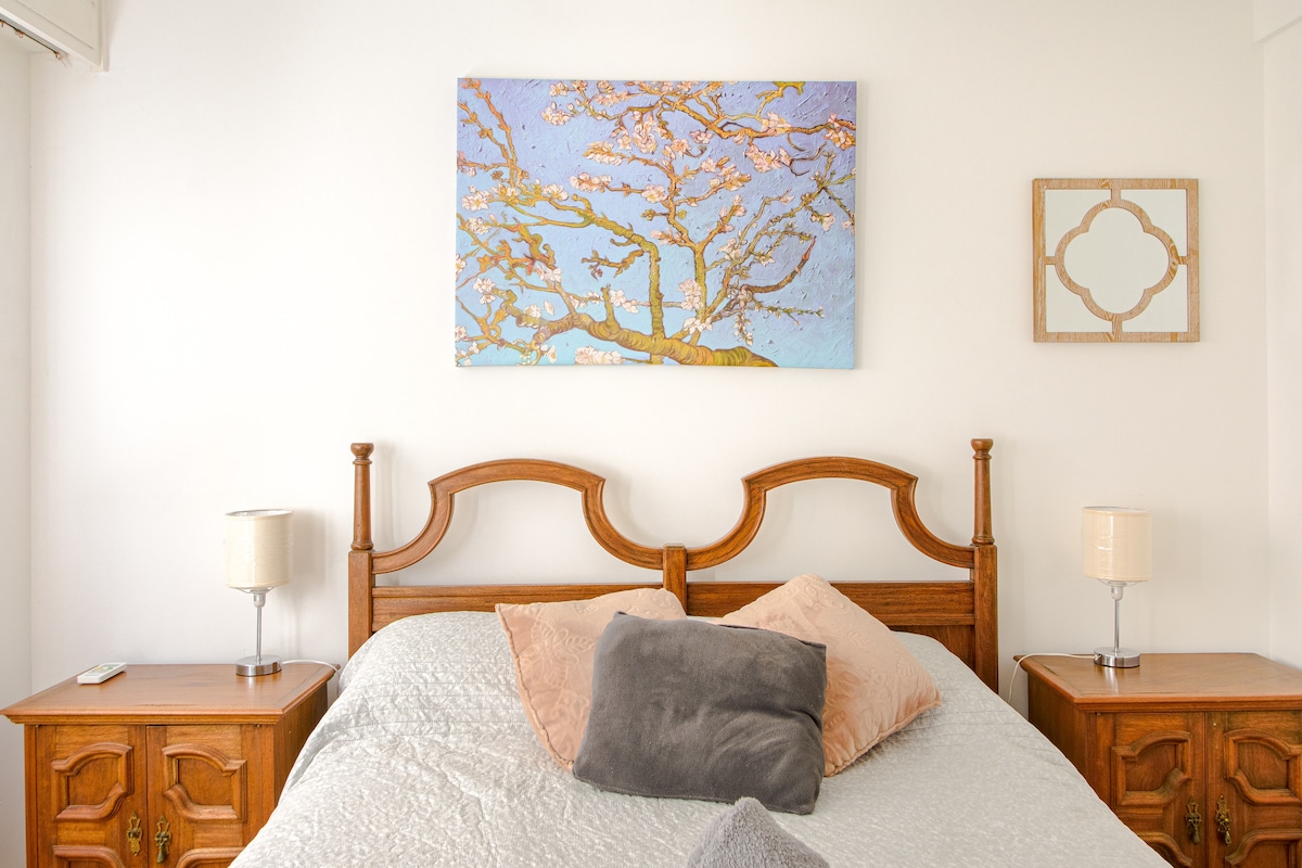 The bedroom features a wooden bed frame with a light-colored bedding set. An abstract painting of a tree is displayed above the headboard. Wooden nightstands with lamps are positioned on either side of the bed, contributing to a neutral and serene environment.