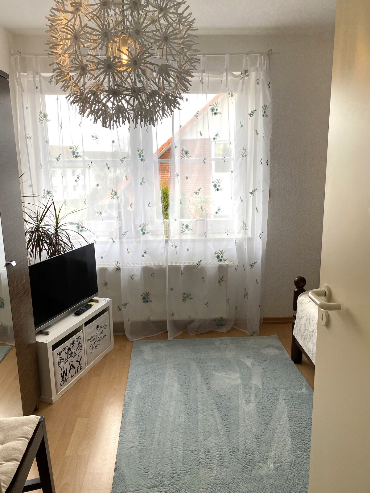 A well-lit room is presented, featuring a large window with sheer floral curtains that allow natural light to filter in. A decorative light fixture hangs from the ceiling, and a soft rug lies on the floor. A small television and a comfortable chair are also visible.