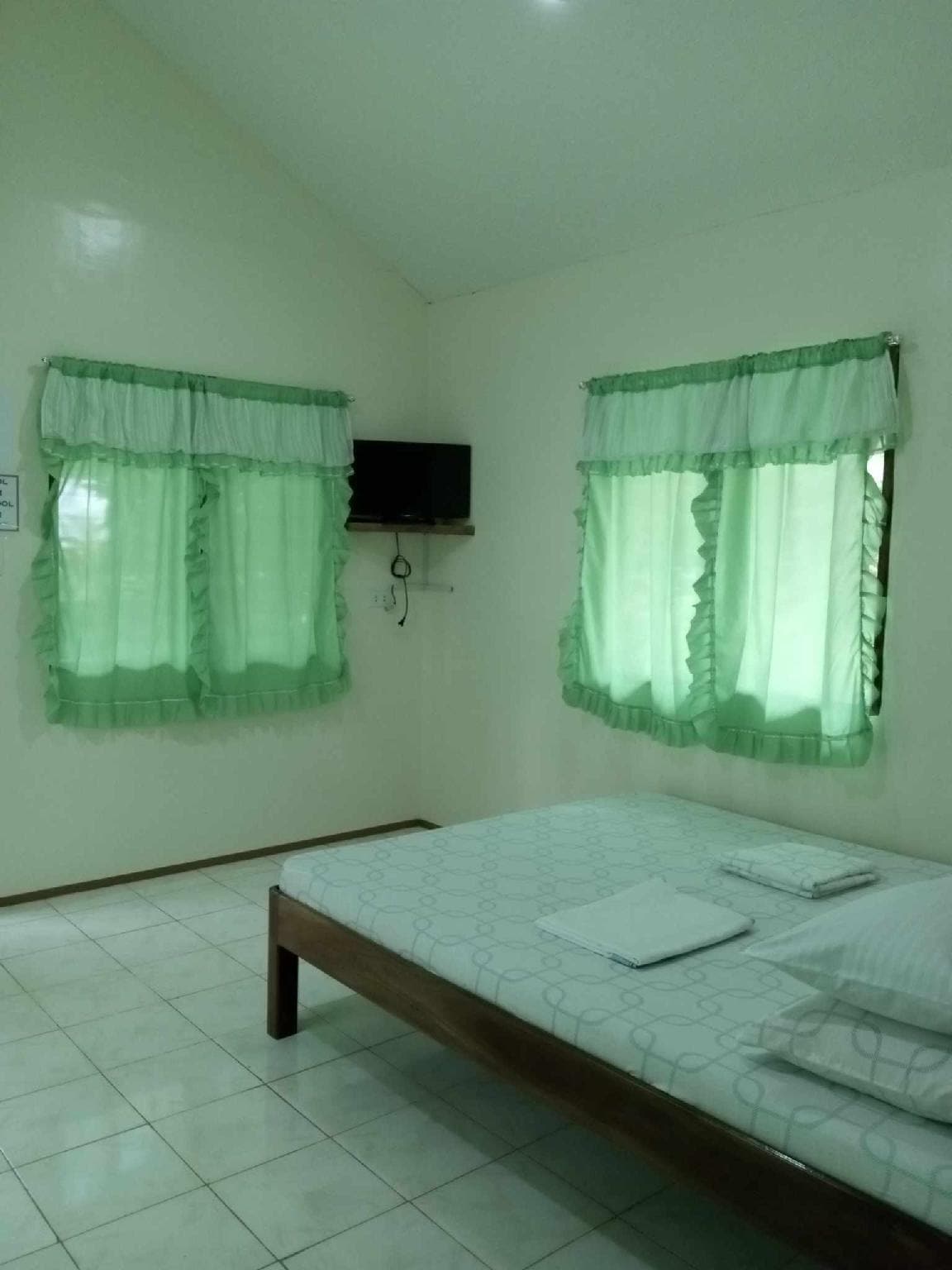 A simple bedroom is featured with a spacious bed covered in white linens, complemented by two large windows adorned with green curtains. A small television is mounted on the wall, enhancing the room's functionality. Neutral walls and tiled flooring contribute to a bright atmosphere.