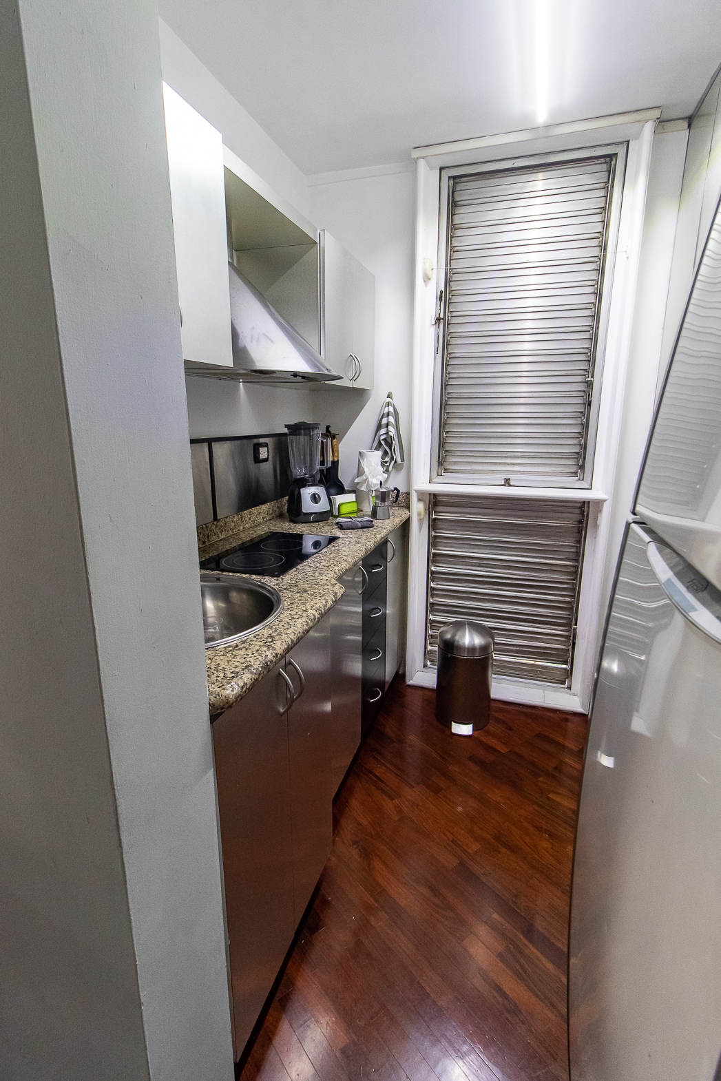 A compact kitchen is presented, featuring stainless steel cabinets and appliances. A granite countertop borders a double sink and stove. Natural light enters through a window, with a metallic trash bin positioned nearby. Smooth wooden flooring enhances the overall clean and modern appearance.