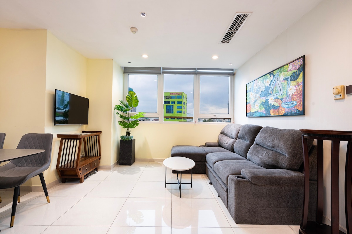 A spacious living area features large windows allowing natural light to illuminate the space. A comfortable gray sectional sofa is positioned near a round coffee table. A flat-screen TV is mounted on the wall, complemented by a decorative plant and an artwork above a wooden console.