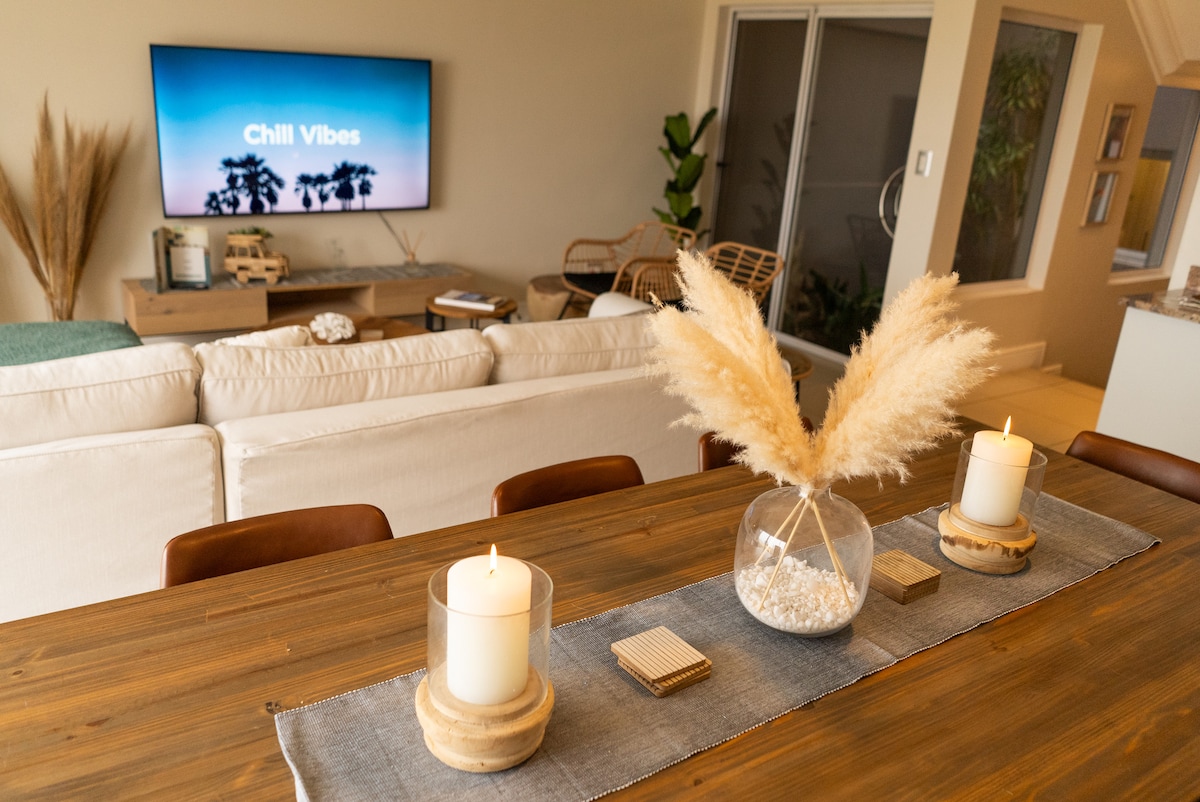 A dining area features a wooden table adorned with a vase of dried pampas grass and two candles. In the background, a comfortable seating arrangement is visible, alongside a television displaying the words 'Chill Vibes' and a plant by the window.