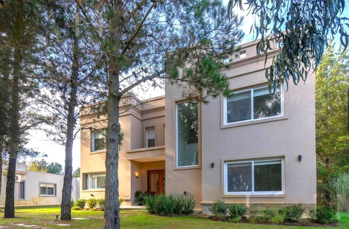 A modern two-story house is surrounded by tall trees and a manicured lawn. Large windows allow natural light to fill the interior, showcasing a welcoming entrance. The neutral exterior is complemented by a simple landscape design, enhancing the overall spacious appearance.