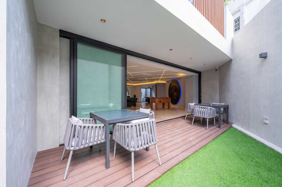 An outdoor terrace features two modern dining tables surrounded by matching chairs. The space is complemented by a green lawn, with large sliding glass doors leading into a contemporary living area, offering a seamless transition between indoor and outdoor relaxation.