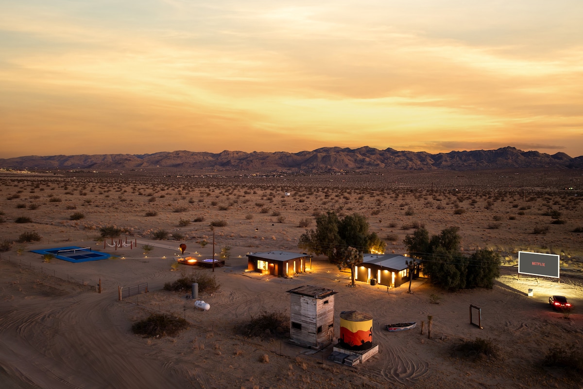 An aerial view captures a private desert compound under a colorful sunset, with amenities visible including a swimming pool, drive-in movie screen, and nearby structures. The surrounding landscape features open desert terrain with distant mountains, enhancing the remote and peaceful setting.