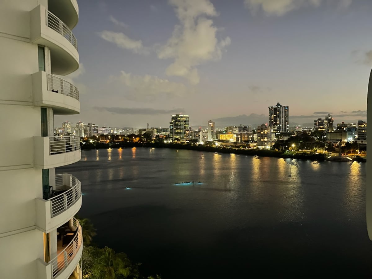 A panoramic view of the lagoon and cityscape during twilight, featuring twinkling lights reflecting on the water's surface. The scene captures a blend of modern buildings and lush greenery, framed by the gentle curves of the building’s balcony. Soft pastel hues grace the sky.