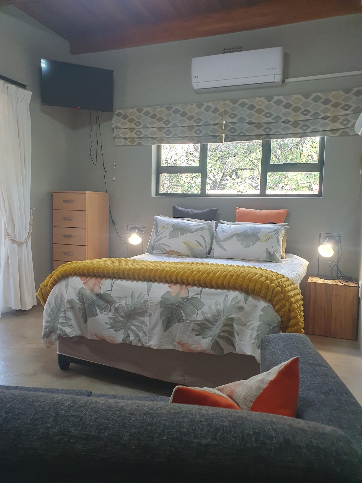 A cozy bedroom is furnished with a double/queen bed adorned with floral bedding and a mustard yellow throw. A television is mounted on the wall, and windows allow natural light to fill the space. A comfortable couch complements the room's relaxed atmosphere.