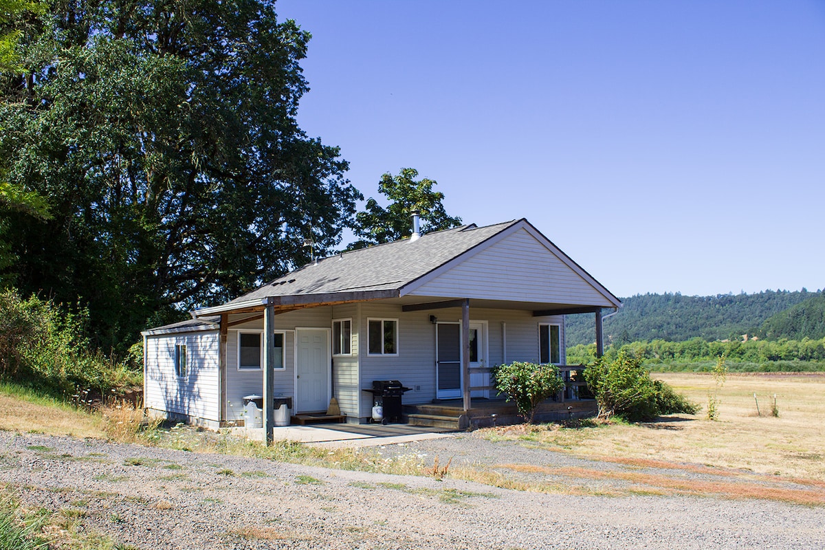 The cottage is situated in an open field, framed by tall trees. The exterior features a covered porch with two entry points, and large windows that provide natural light. Surrounding landscapes include rolling hills and grasslands under a clear blue sky.