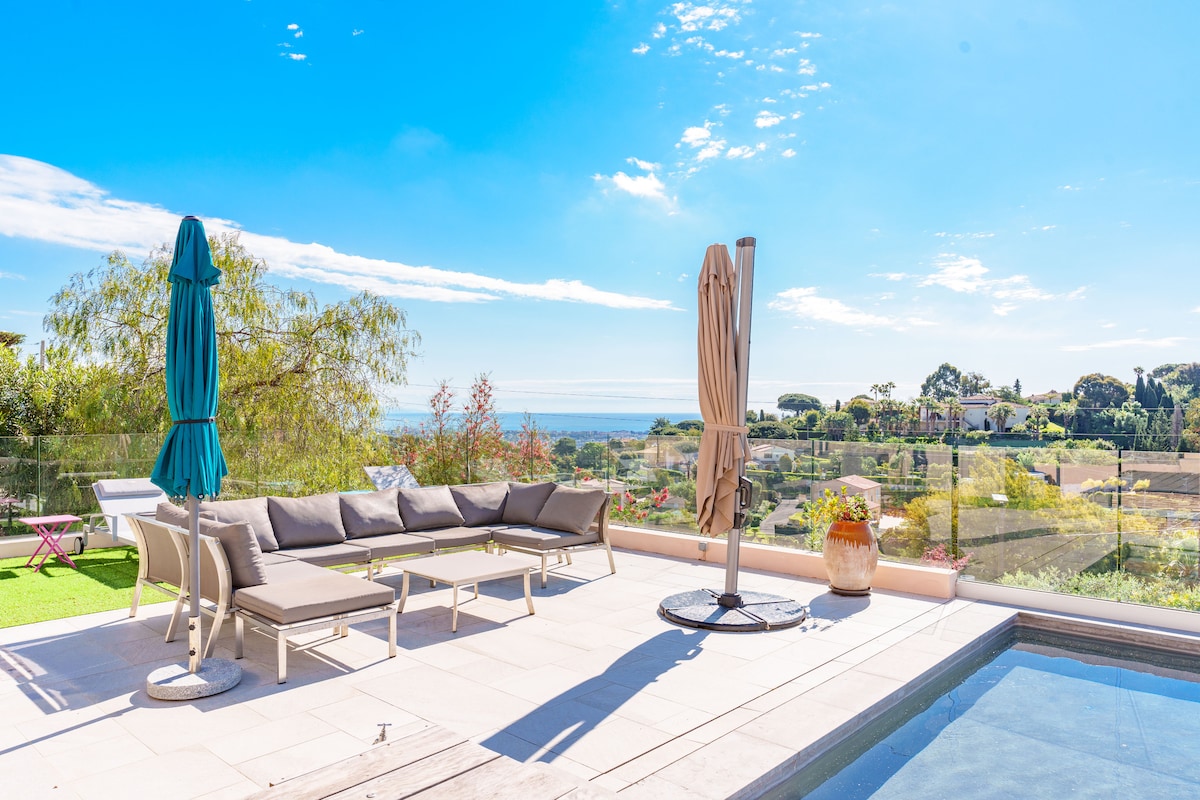 An outdoor terrace is presented with a spacious lounge area featuring gray seating and parasols. A clear view of the sea and landscaped gardens is visible in the background, complemented by a hint of blue sky and scattered clouds.