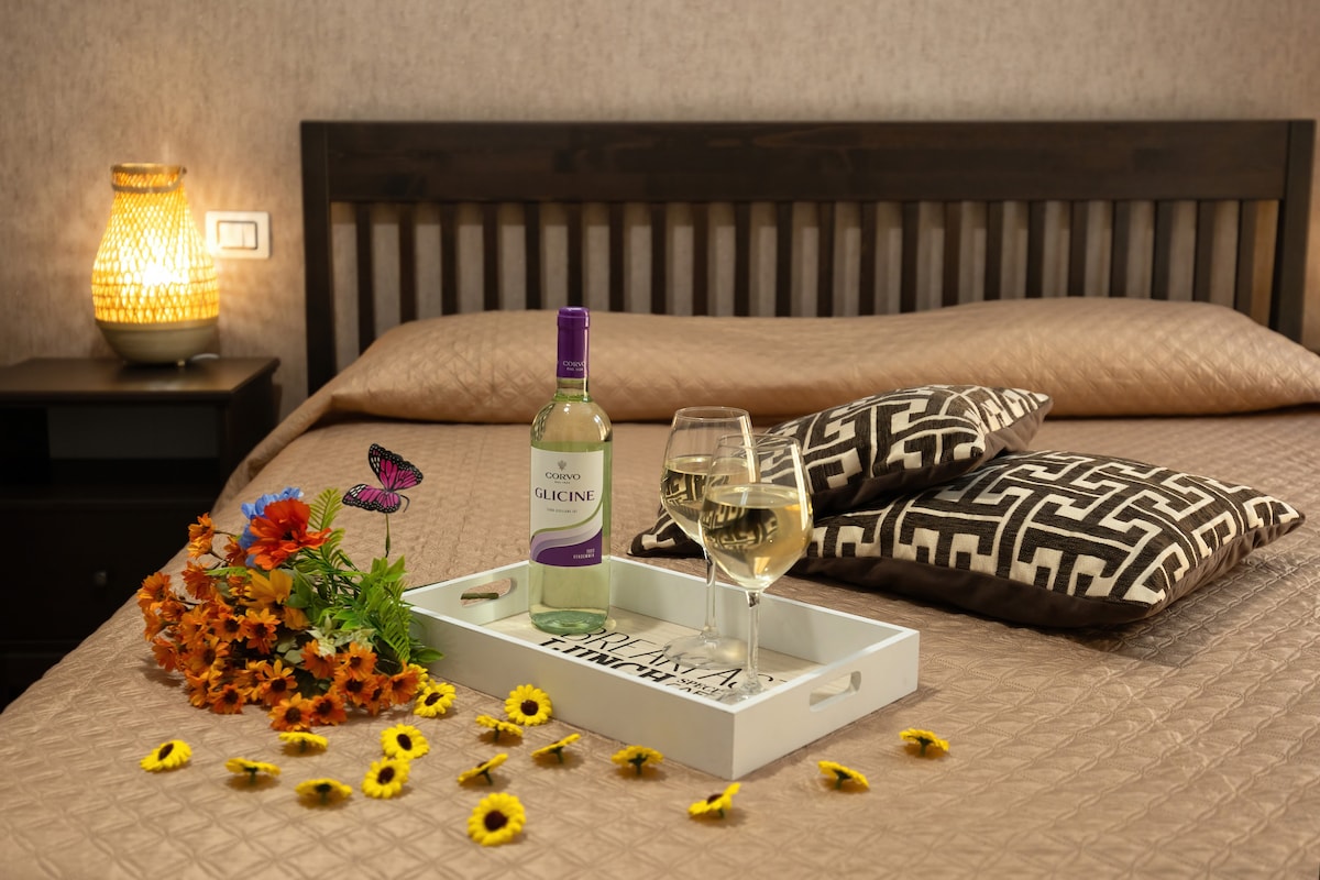 A spacious bed is adorned with a textured beige coverlet and decorative pillows. A tray with a bottle of white wine and a glass sits atop the bed, accompanied by a vibrant arrangement of flowers. A soft light is provided by a nearby lamp.