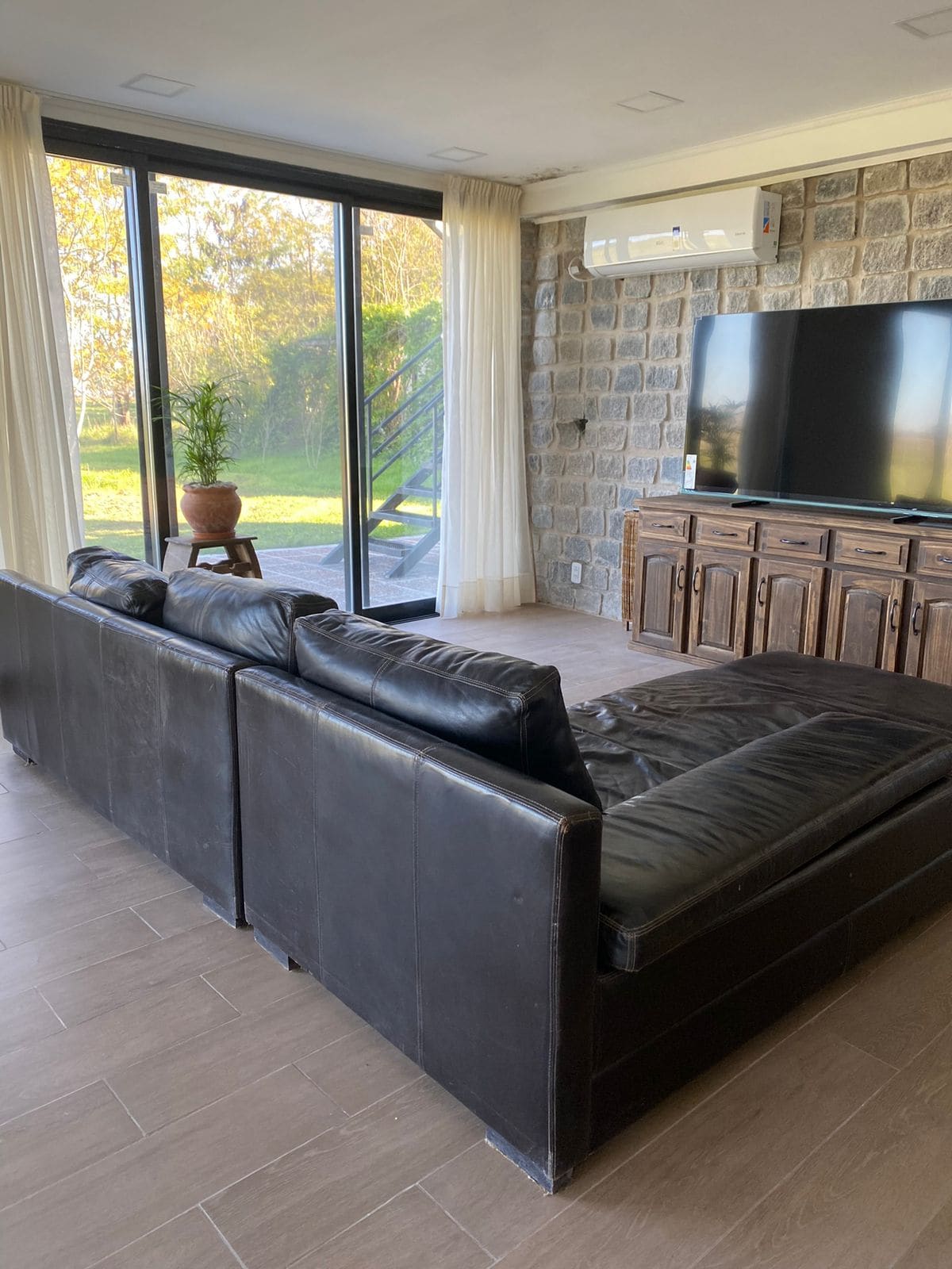 A spacious living area features a large black sectional sofa positioned near a wall of windows, allowing natural light to illuminate the room. A wooden media console sits against a stone wall, and a potted plant adds a touch of greenery in the corner.