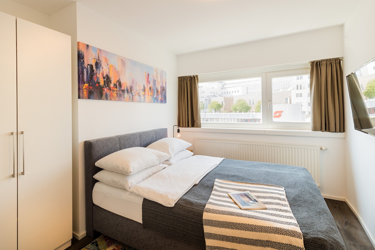 A modern bedroom features a spacious bed with crisp white linens and a textured gray blanket. Large windows offer ample natural light, complemented by curtains. A piece of abstract artwork adds a pop of color to the white walls, enhancing the room's contemporary design.