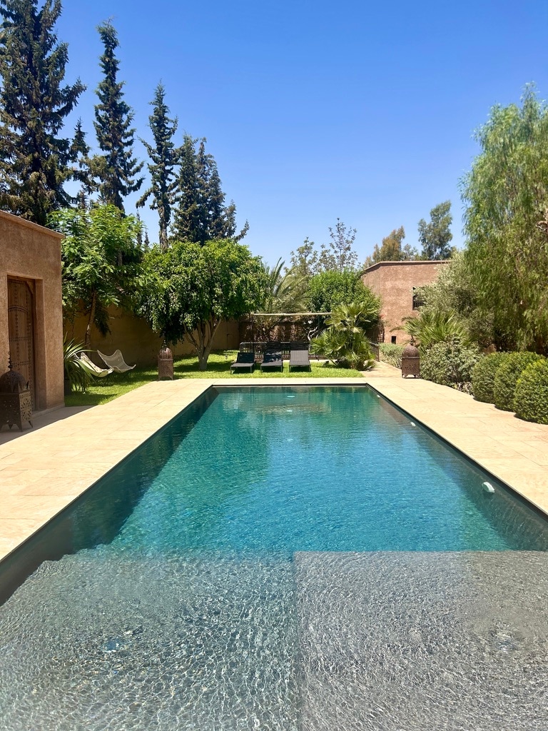 A tranquil swimming pool with clear blue water reflects the surrounding greenery. Sun loungers are positioned nearby, and tall trees provide shade along the perimeter. The garden features neatly trimmed hedges and a seating area in the background, enhancing the peaceful atmosphere.