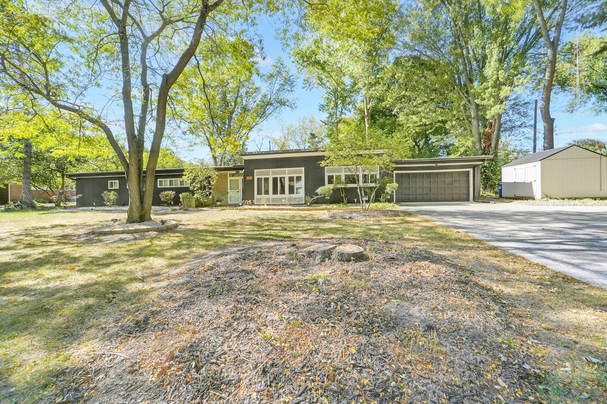 A spacious modern home is set amidst lush greenery, featuring a large front yard with scattered rocks and trees. The driveway leads to a two-car garage, and the house is accented by ample natural light through its expansive windows.