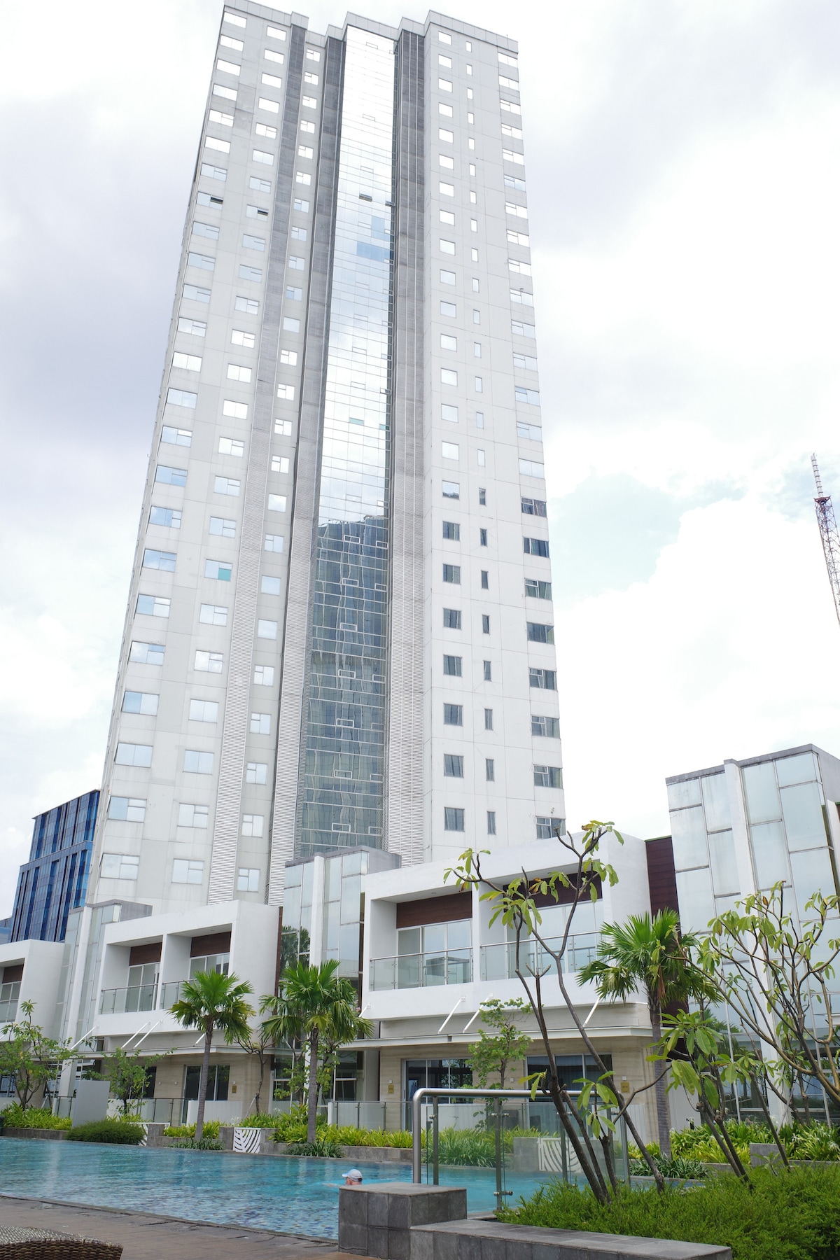 A modern high-rise building is depicted, featuring a sleek glass facade that reflects the surrounding greenery. The lower levels showcase a landscaped area with palm trees and a swimming pool, providing a refreshing outdoor space next to the structure.