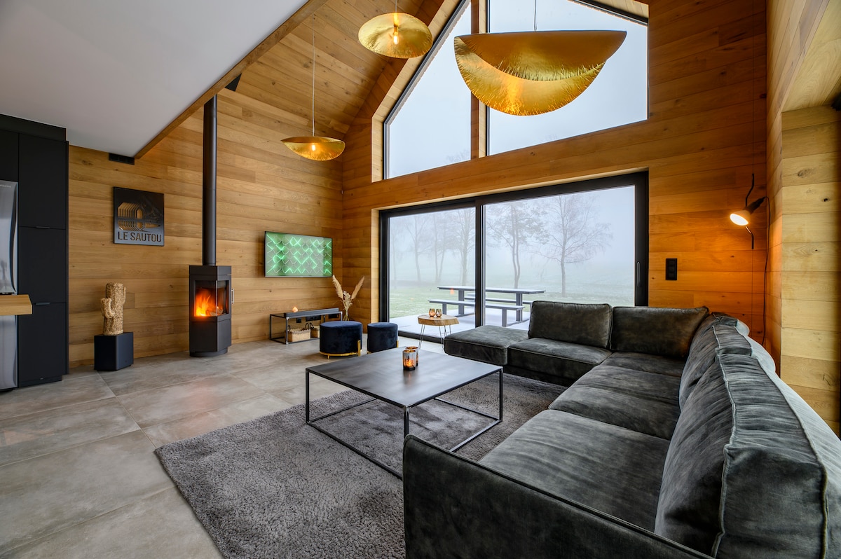 A spacious living area features a large sectional sofa, a coffee table, and a sleek fireplace. Floor-to-ceiling windows allow natural light, creating a connection to the outdoors. The wooden walls add warmth, while modern light fixtures hang from the vaulted ceiling.