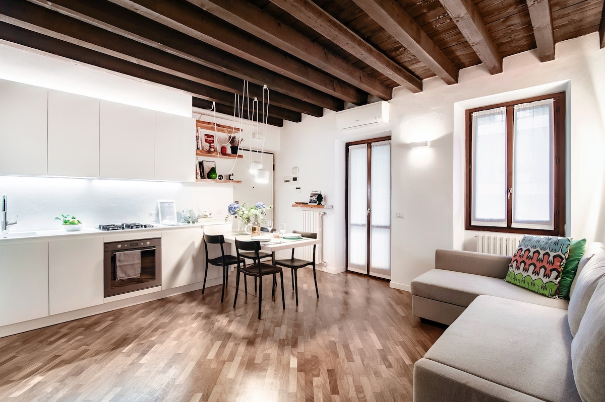 The living area features wooden beams overhead and a light wooden floor. A fully equipped kitchen is visible, accompanied by a dining table with four chairs. A double-size sofa bed is positioned nearby, with natural light entering through a window beside the entrance.