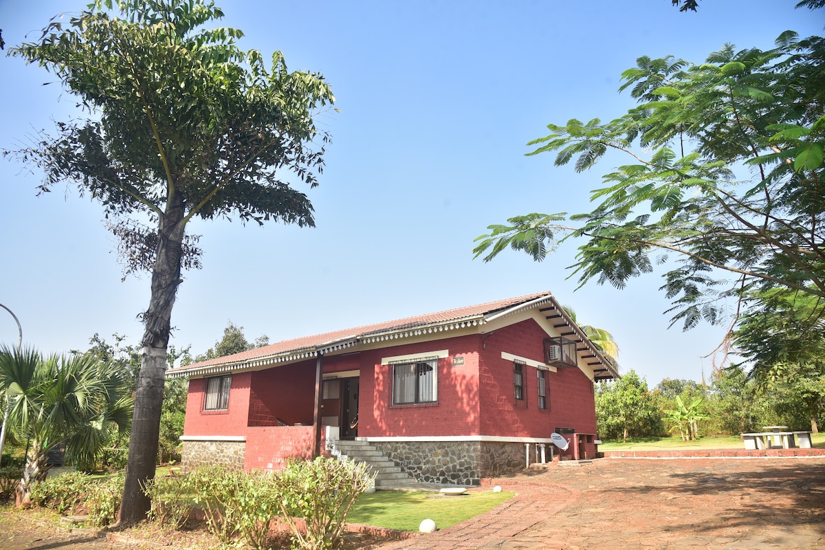 A single-story villa is presented in a vibrant red hue, surrounded by lush greenery and tall trees. The entrance features a solid stone foundation, and a clear blue sky enhances the natural setting. Well-maintained gardens frame the property, offering a serene view.