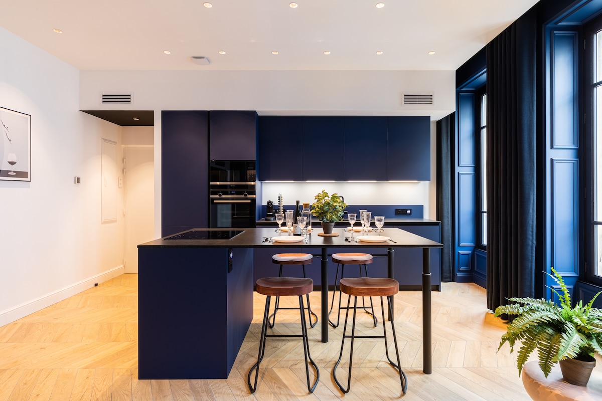 A modern kitchen displays sleek dark cabinetry and a contrasting light countertop. A central island is equipped with seating for two and is set with diningware. Sunlight filters through large windows adorned with dark curtains, highlighting the warm wooden flooring.