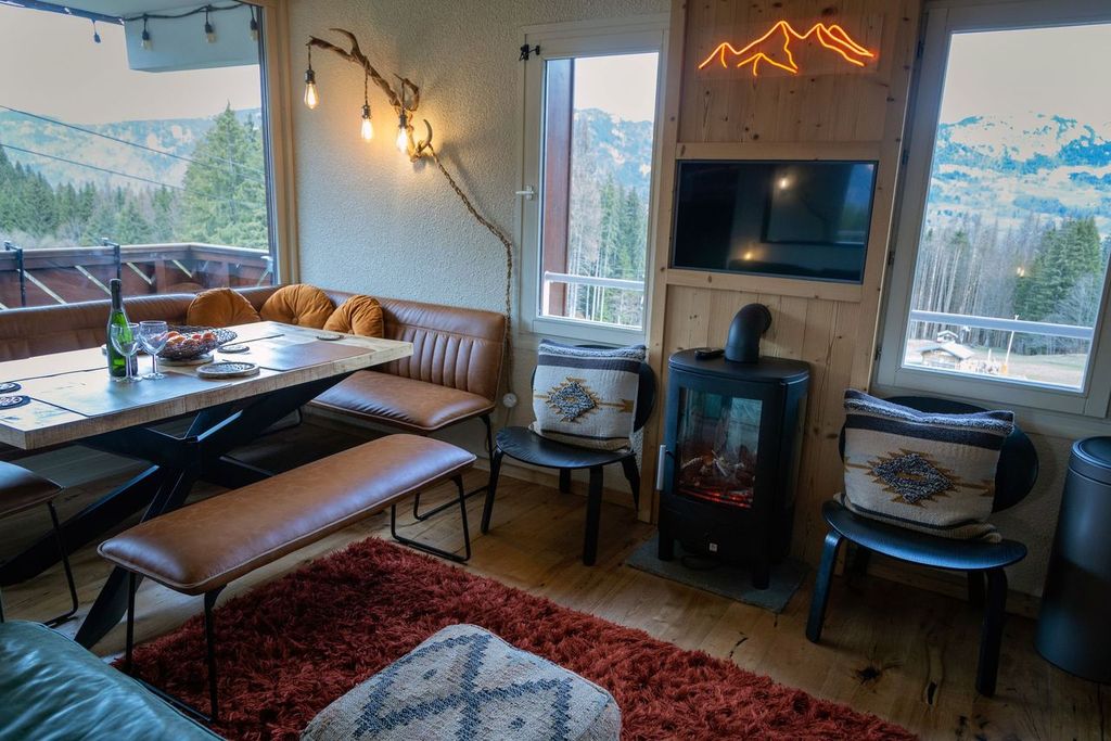 A cozy living area features a modern dining table, leather seating, and a wall-mounted TV. An electric fireplace provides warmth, while large windows reveal stunning mountain views. Decorative pillows add comfort, and soft lighting enhances the space's inviting feel.