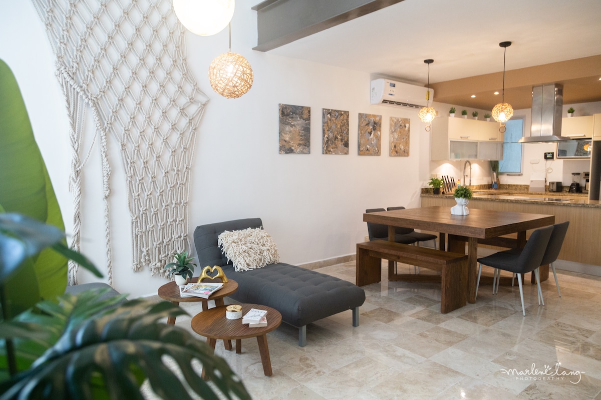 A stylish living area features a comfortable sectional with a textured throw pillow and a decorative coffee table. Artwork hangs above a spacious dining table surrounded by four chairs. Modern lighting fixtures cast a soft glow, while greenery adds freshness to the space.