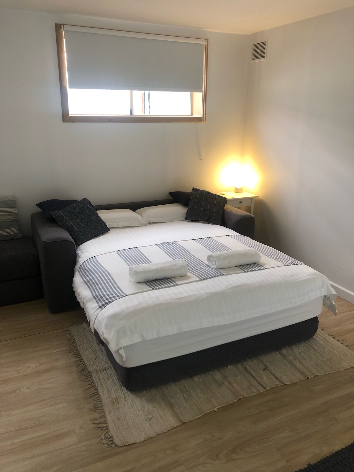 A cozy sleeping area is designed with a spacious bed featuring crisp white linens and two rolled towels placed on top. A soft throw blanket adds texture, while a small bedside lamp emits gentle light beside a window, enhancing the inviting space.