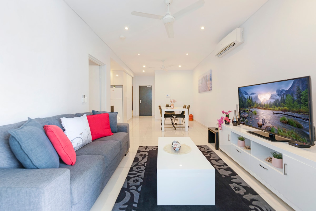 A modern living room features a comfortable gray sofa adorned with red accent pillows. A sleek coffee table sits on a decorative rug, and a large flat-screen TV displays a scenic image. The space is brightened by natural light and includes a dining area visible in the background.