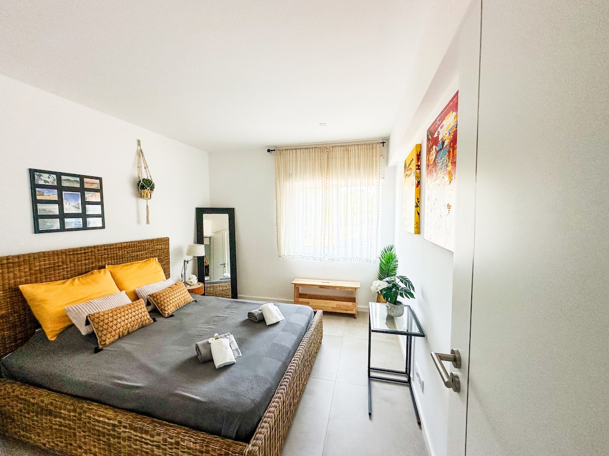 A well-lit bedroom features a spacious bed adorned with yellow and textured pillows. A small table beside the bed holds a lamp, while a full-length mirror reflects the room. Natural light filters through a window, enhancing the room's neutral decor.