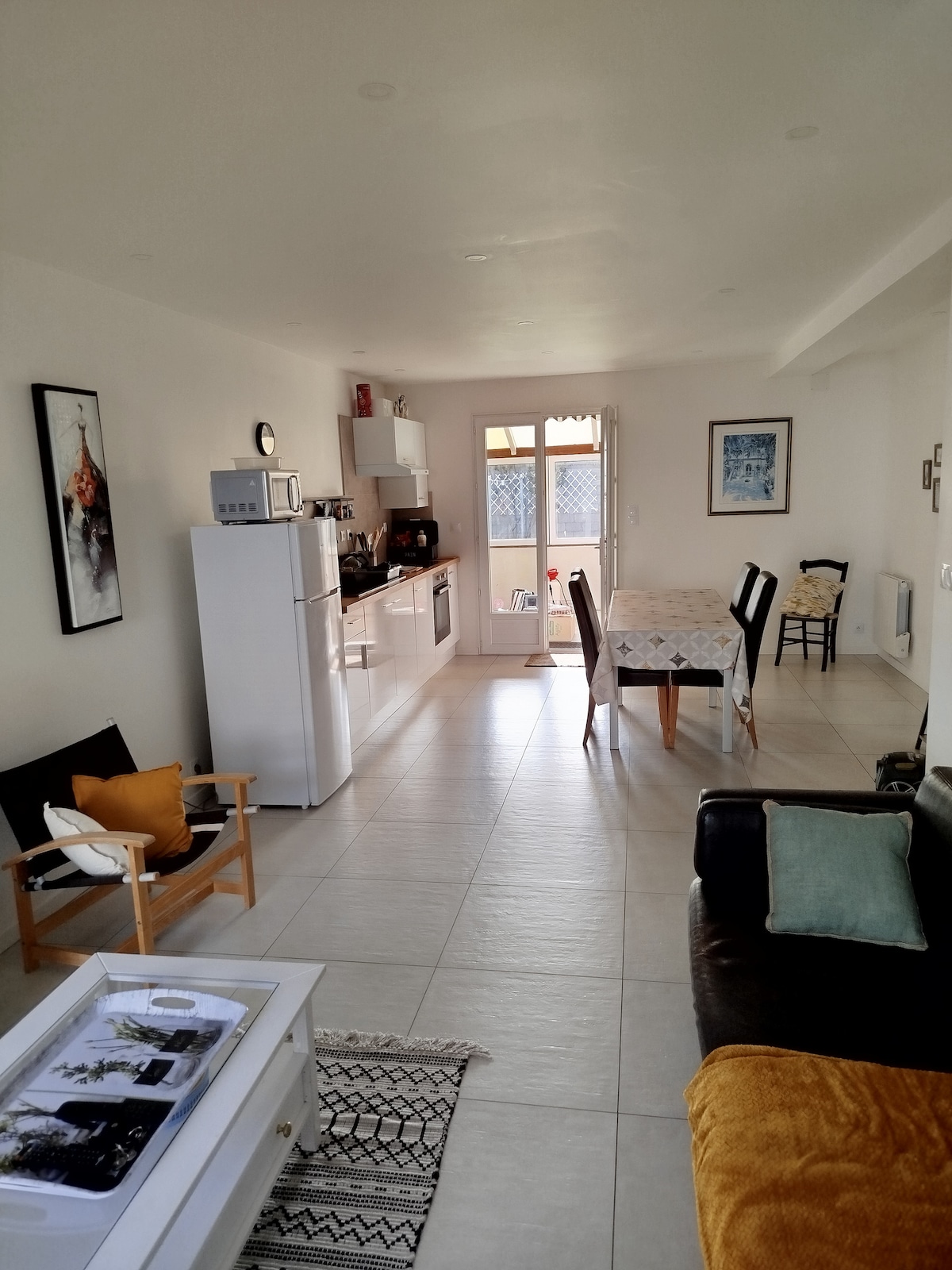 A bright living area features a white dining table with patterned linens, complemented by a black chair. A spacious kitchen is equipped with modern appliances, including a white refrigerator. Light enters through a glass door leading to an outdoor space, while cozy seating arrangements are present.