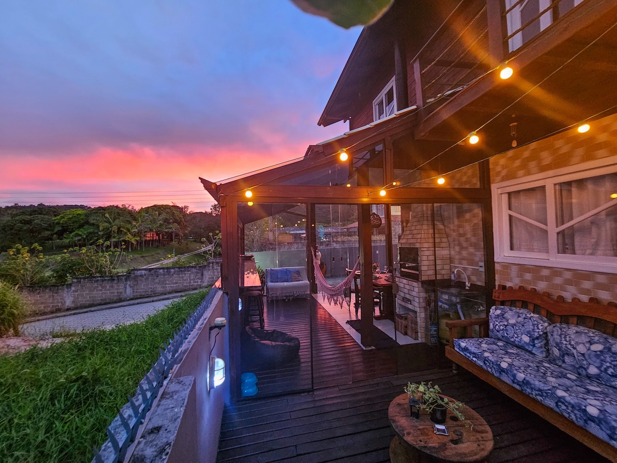 An outdoor deck is illuminated by warm string lights, creating a cozy atmosphere. A comfortable seating area with patterned cushions overlooks a lush garden. A vibrant sunset casts soft colors across the sky, enhancing the natural beauty of the surroundings.