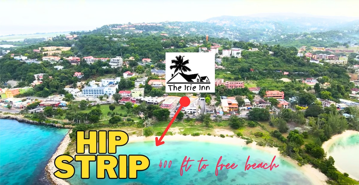 An aerial view showcases the vibrant surroundings of The Irie Inn, situated on the hip strip. The coastline and lush green hills are visible, highlighting the proximity to a free public beach. Notable landmarks and colorful buildings punctuate the landscape, creating a lively atmosphere.