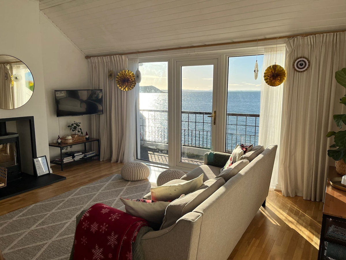 A spacious living area is depicted with large glass doors revealing views of the sea. Natural light illuminates the room, highlighting a soft grey couch and decorative cushions. A TV is mounted on the wall, and a cozy rug covers the wooden floor.