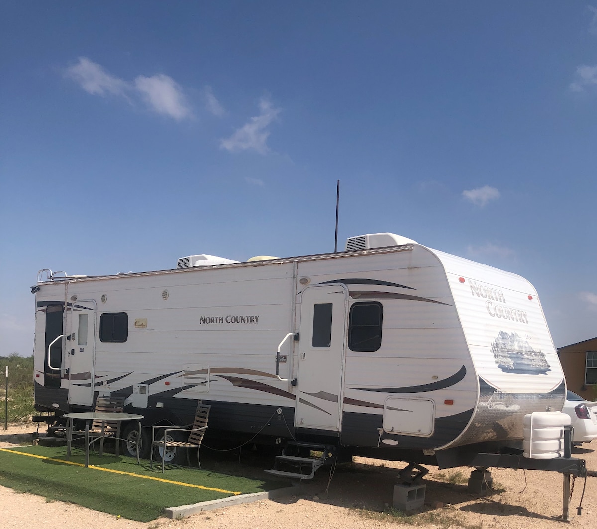 A spacious RV is shown parked on a gravel surface, displaying a white exterior with dark accents. The structure is fitted with multiple windows and features steps leading up to the entrance. A clear blue sky provides a bright backdrop.