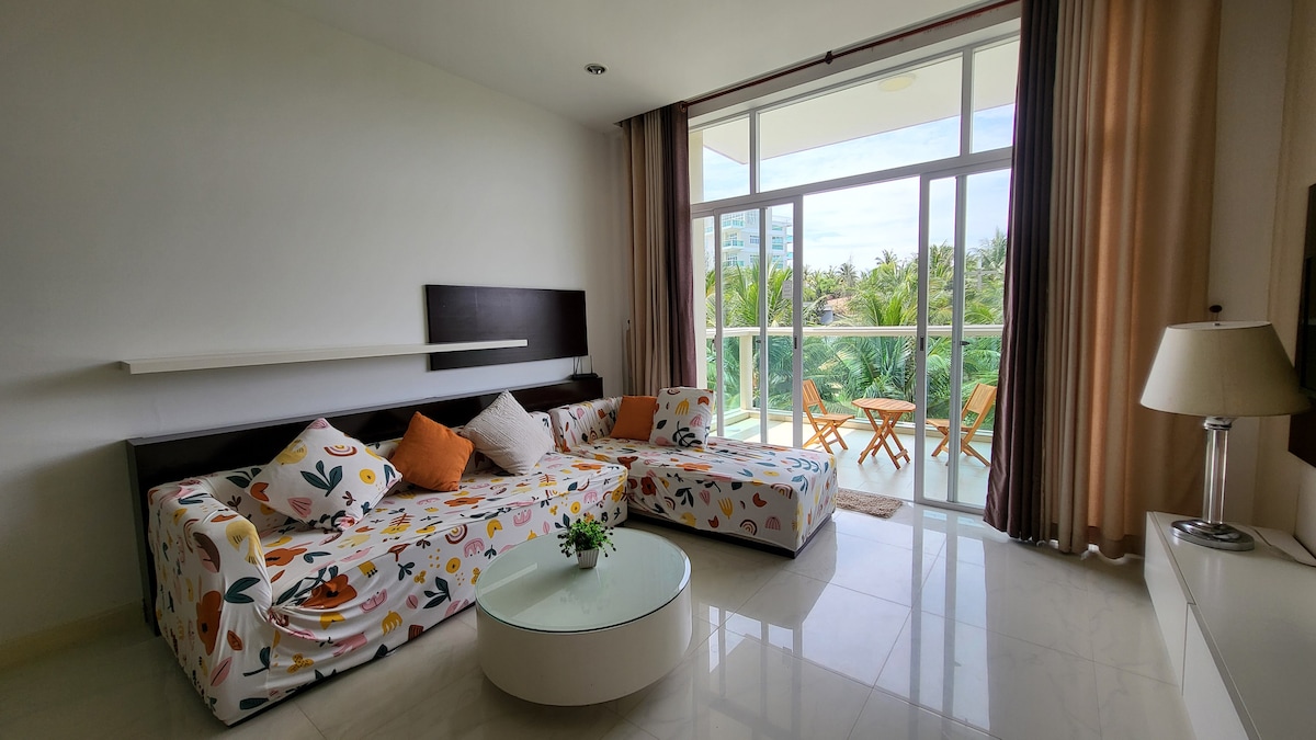 A bright living area features a large, comfortable lounge sofa adorned with a colorful pattern. A small round coffee table sits at the center, while a pair of outdoor chairs and a table can be seen through the balcony doors, overlooking lush greenery.