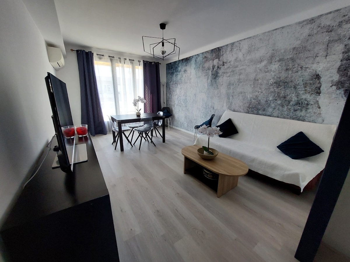A living area is presented with a sleek design featuring light wood flooring and a textured wall. A comfortable sofa is accompanied by a wooden coffee table. Natural light filters through large windows, and a dining table is set for four, enhancing the inviting atmosphere.