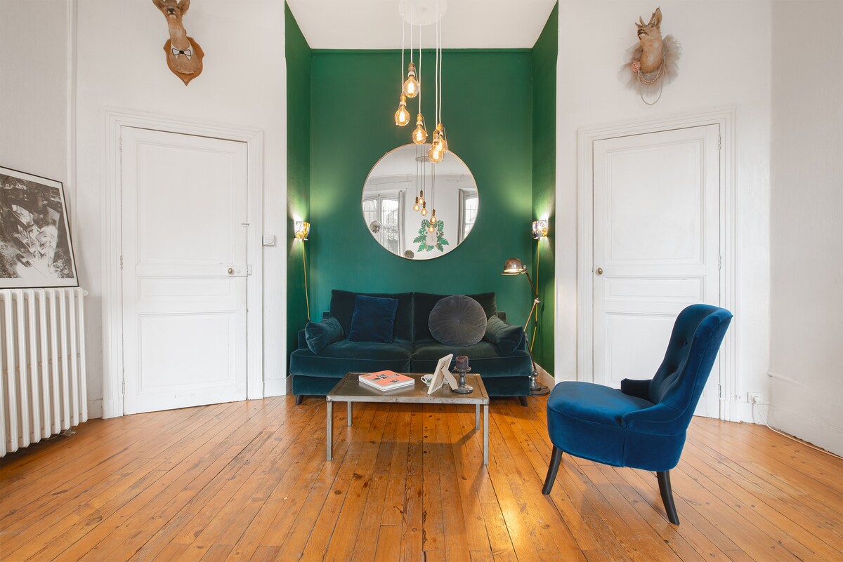 A cozy living area is presented with a green accent wall and wooden flooring. A dark blue sofa is paired with a velvet armchair, complemented by a modern coffee table. Unique wall art and pendant lights add character to the space, while natural light enhances the atmosphere.