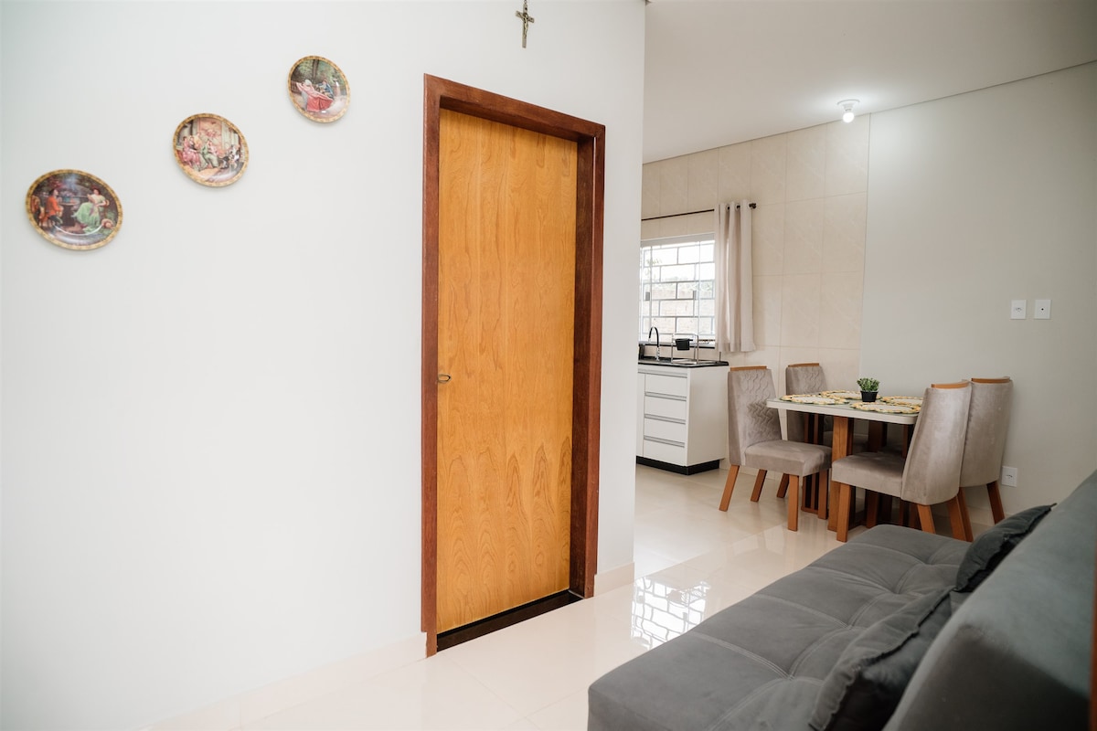 A spacious living area features a comfortable gray sofa, with a wooden door leading into another room. Wall art is displayed on the white wall, and a dining table with upholstered chairs is visible in the background, set against a well-lit kitchen.