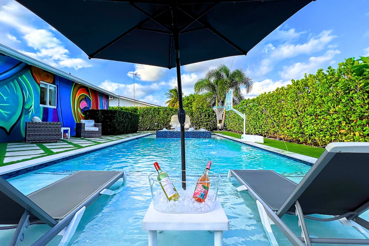 A serene pool area is showcased with two lounge chairs positioned near the water, featuring a small table with bottles resting in ice. The vibrant mural on the nearby wall adds a colorful backdrop, while lush greenery surrounds the outdoor space.