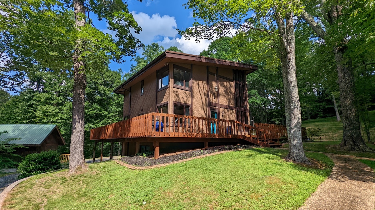 The exterior of the house is set amid lush greenery, featuring large windows that provide ample natural light. A wraparound deck with wooden railings offers outdoor space for relaxation, while the sloping landscape creates a serene atmosphere in the surrounding wooded area.