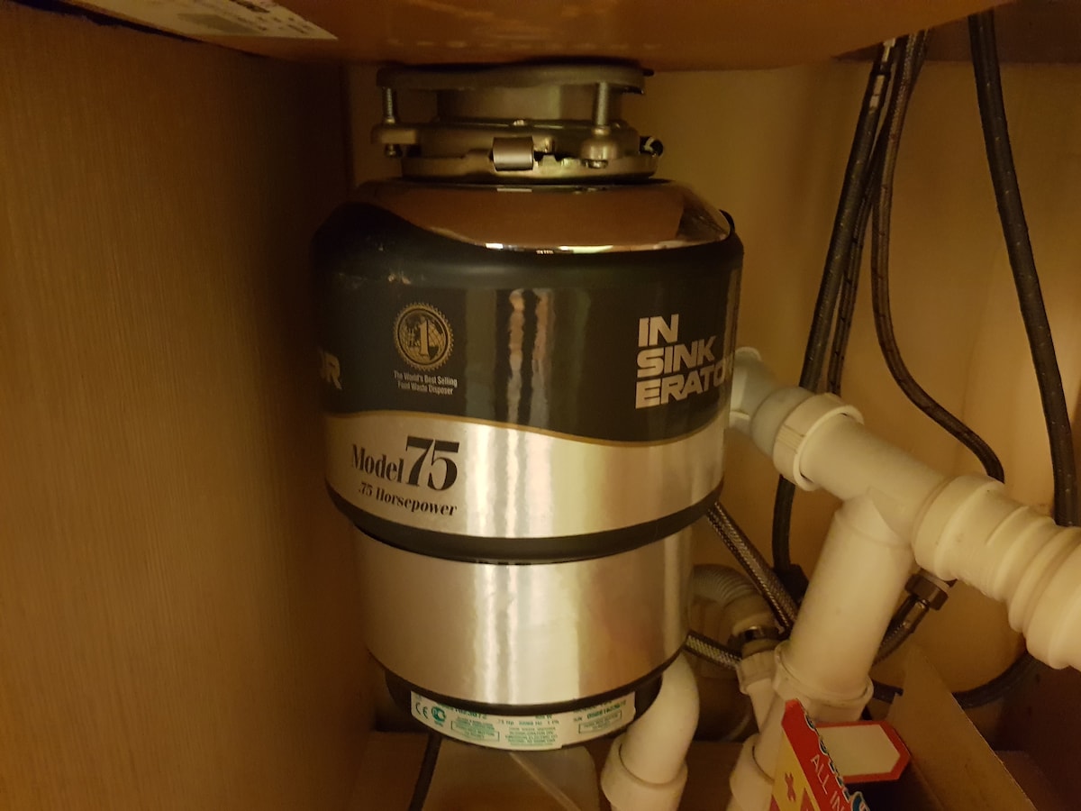 An InSinkErator garbage disposal unit is installed beneath a kitchen sink, featuring a stainless-steel finish. Various plumbing and electrical connections are visible, providing a functional aspect to the kitchen's waste management system.