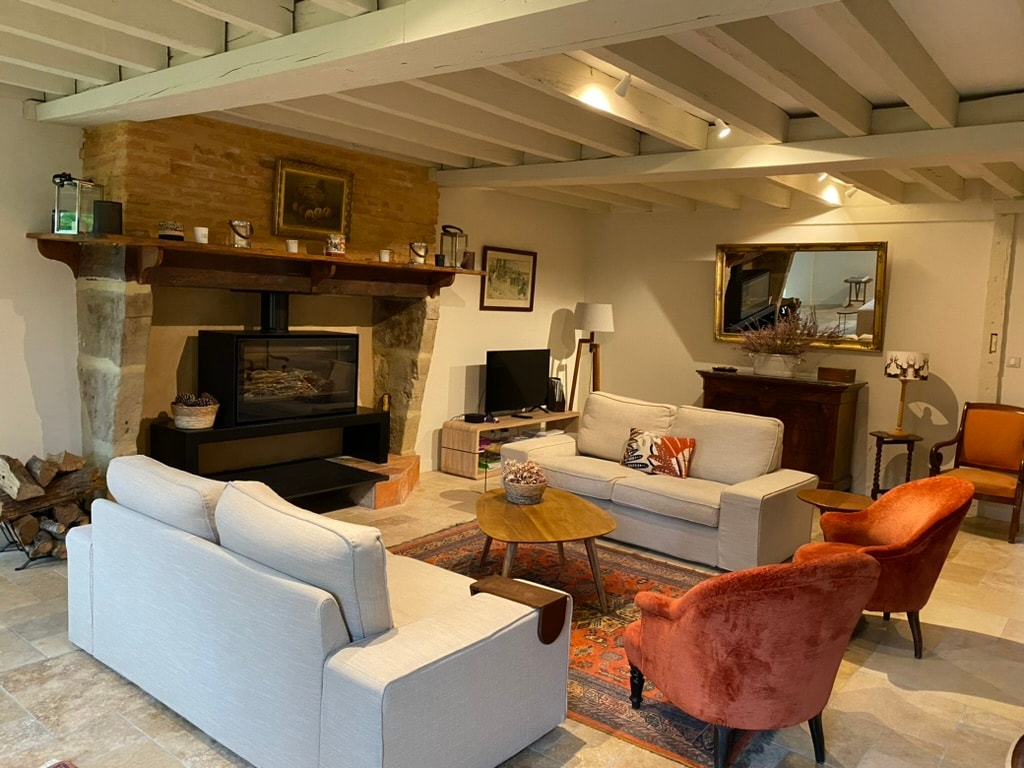 A spacious living area is furnished with contemporary seating arrangements, including a light-colored sofa and orange-accented chairs. A rustic fireplace is set against a stone wall, while a flat-screen television is positioned nearby. Natural light illuminates the room, highlighting the neutral decor and tasteful artwork.
