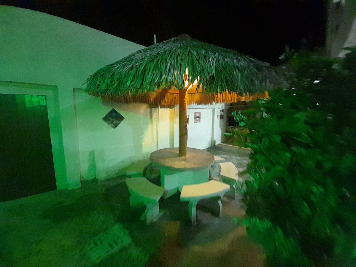 A shaded outdoor seating area is illuminated under soft green lighting, featuring a thatched-roof table with a central wooden post. Surrounding the table, several light-colored chairs offer a comfortable spot for relaxation. Lush greenery frames the space, contributing to a tranquil atmosphere.