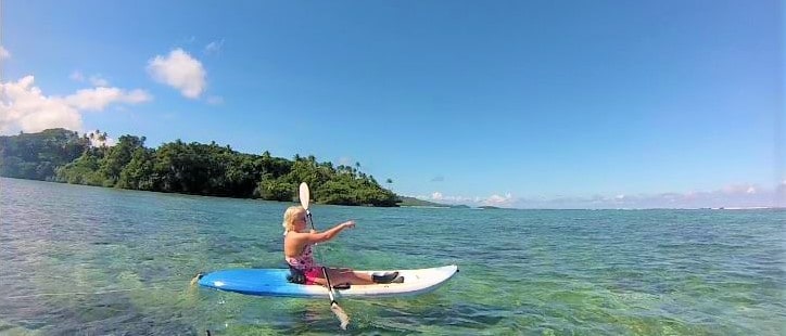 A person is seen kayaking on clear waters, with a vibrant blue sky above and lush greenery in the background. The kayak is partially submerged, showcasing the serene water conditions that allow for easy exploration of the surrounding area.