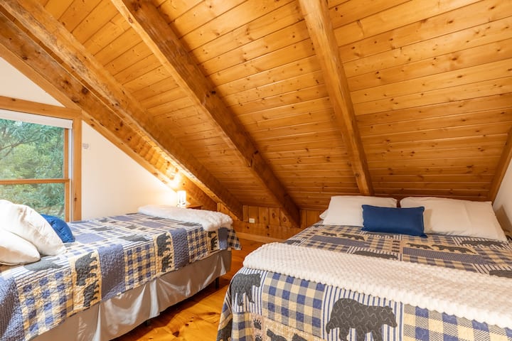 Second-floor bedroom accommodating up to 4 guests with two queen beds. Includes closet, bedside lighting, window, and floor fan for circulation.
