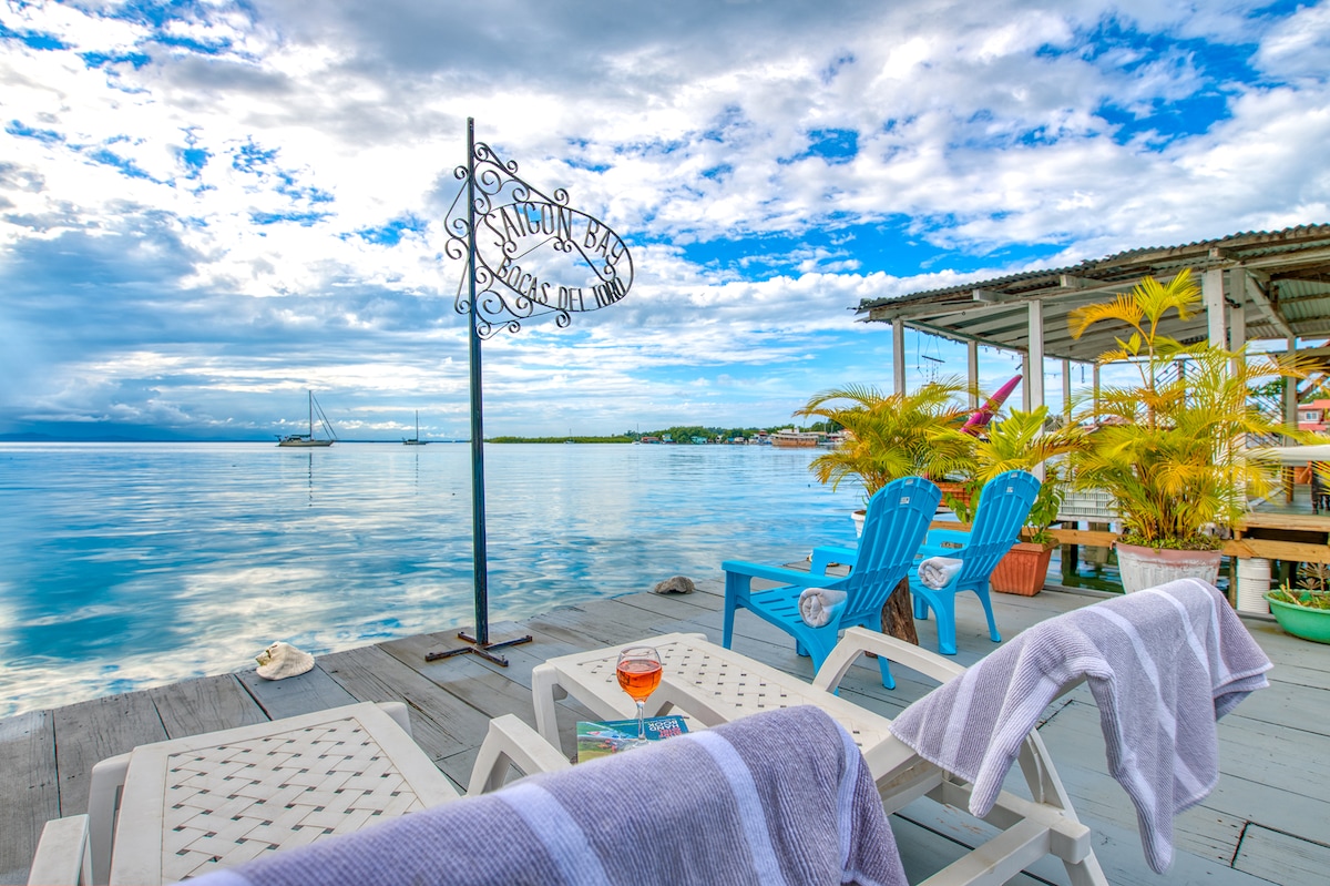 A serene outdoor seating area features lounge chairs arranged on a wooden deck, overlooking calm waters. A sign reading 'Saigon Bay' is visible, accompanied by tropical plants. The sky showcases an array of clouds, with a sailboat gently floating in the distance.