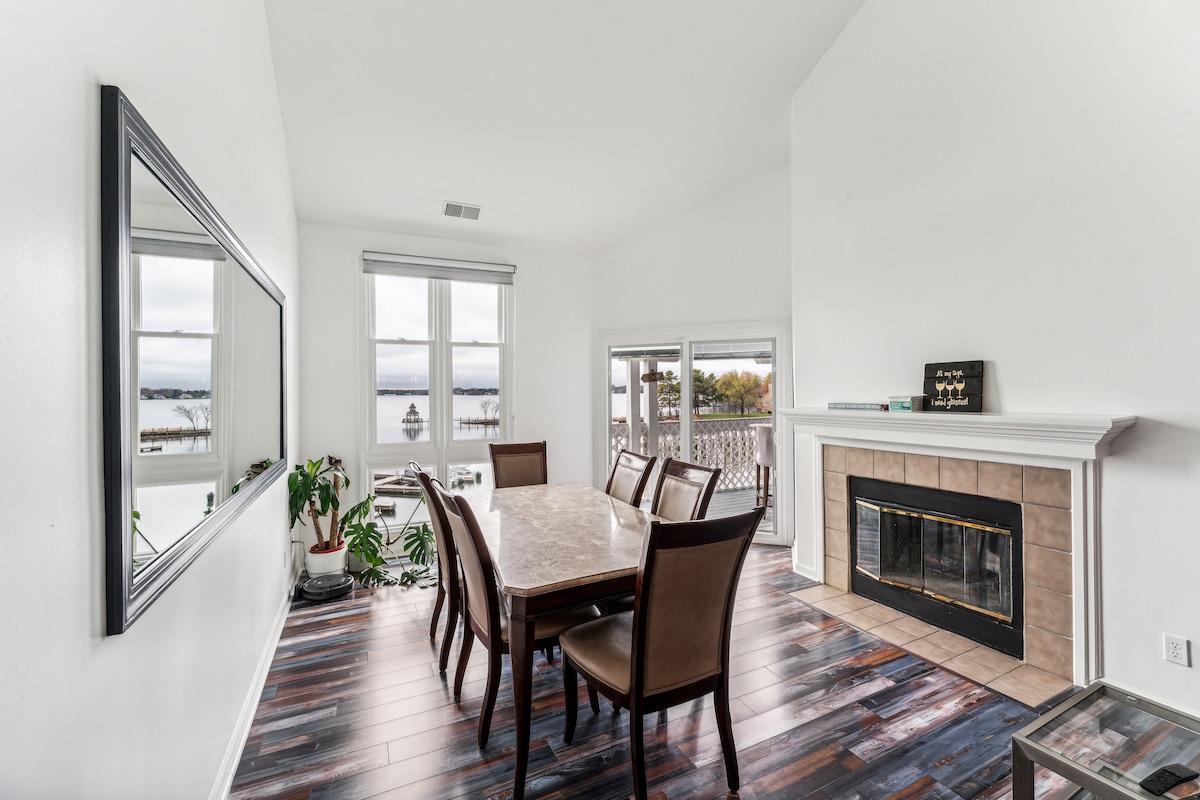 A bright dining area features a large table surrounded by six chairs, positioned near windows that allow natural light to fill the space. A fireplace is located to one side, with a mirror mounted above it reflecting the room's inviting ambiance.